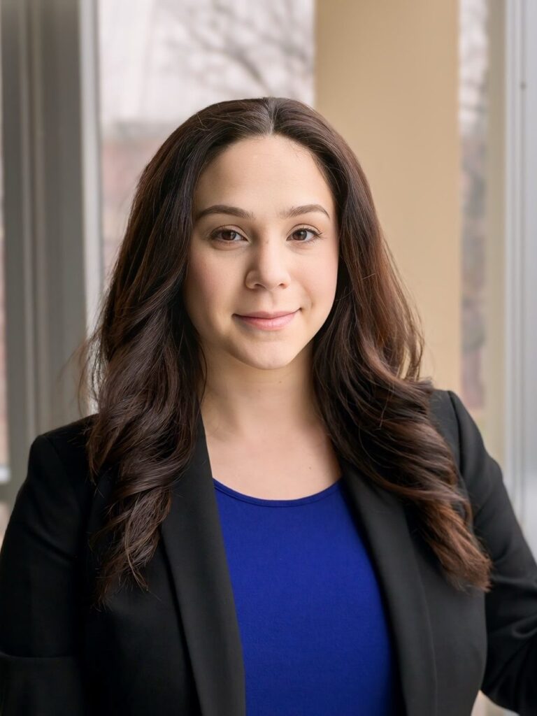 A woman with a blue shirt, brown hair and a black blazer smiles for a portrait inside an academic building.